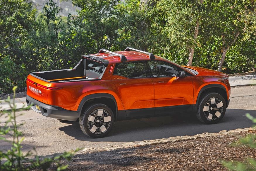 A Fisker Alaska 2023 pickup in burnt orange color is seen driving along a wooded road on a hillside, with trees flanking the road.